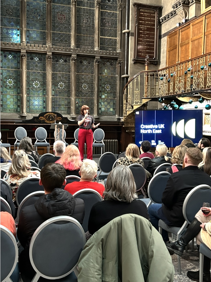 A woman in bright red trousers is holding a microphone and talking to a room full of people. Behind her is a stunning huge leaded windor and the walls are all beautifully lined with wood.